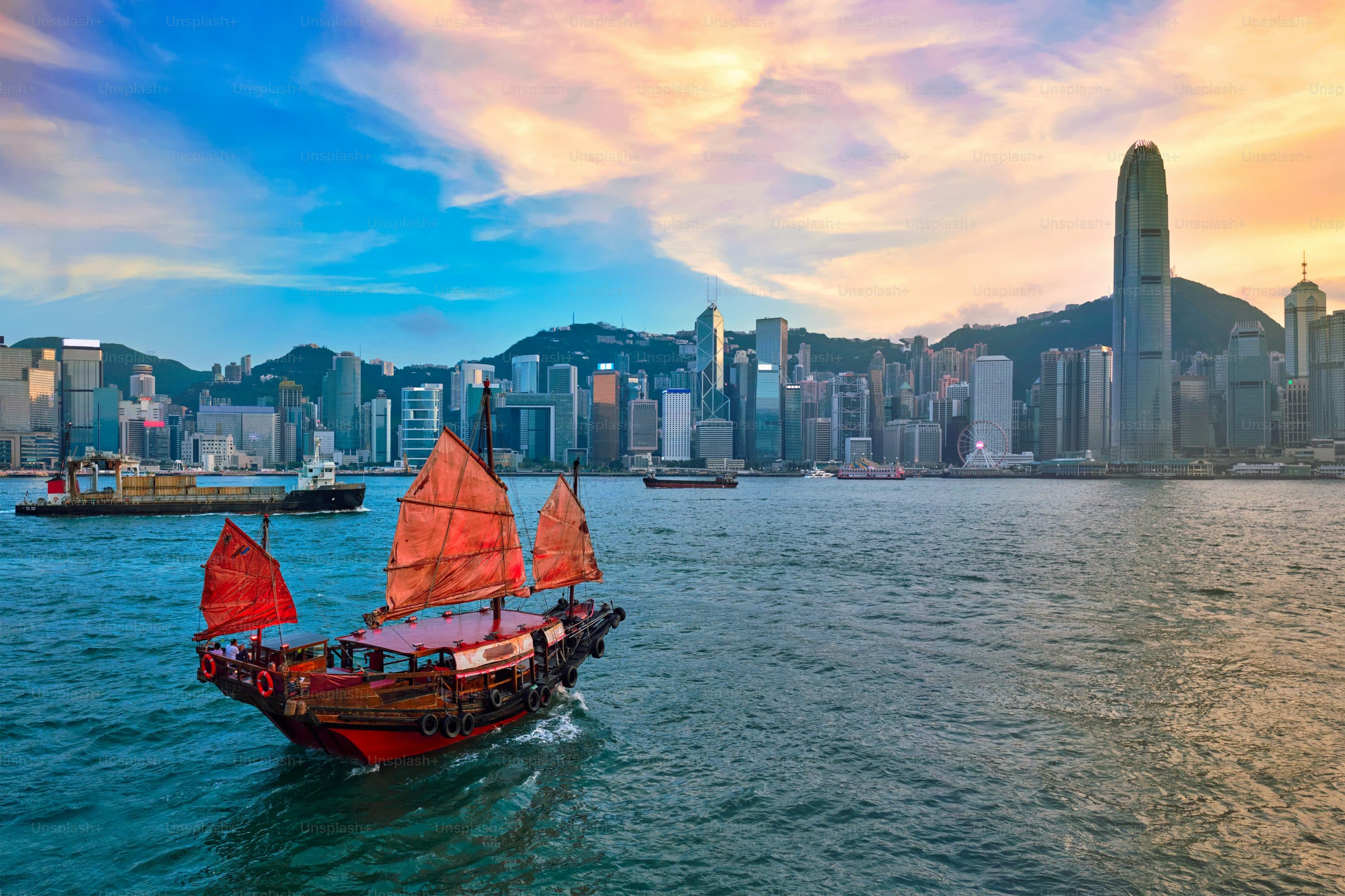 A traditional red-sailed boat crossing Hong Kong harbor at sunset.